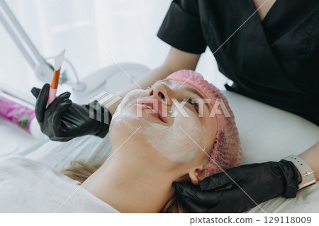 aesthetician doctor applies a creamy face mask on young woman face using a brush aesthetician doctor applies a creamy face mask on young woman face using a brush 129118009