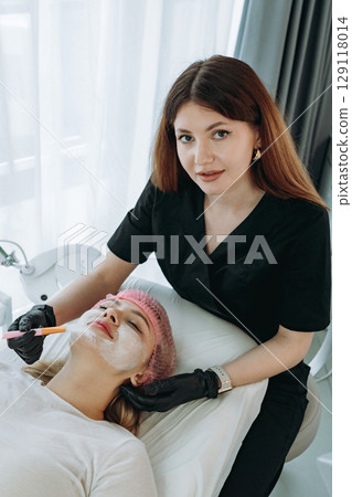 professional aesthetician applies a creamy face mask on young woman face using a brush. Doctor looking at camera 129118014