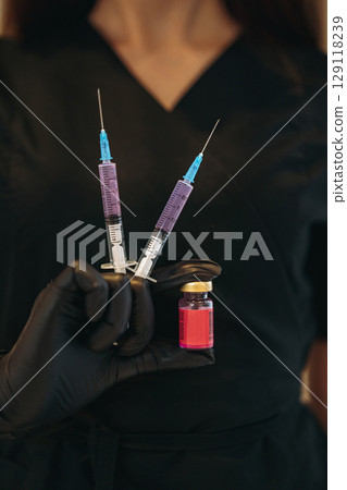 woman hands in gloves wearing black medical suit holding syringes and a nd a small bottle of a cosmetic or medical injectable 129118239