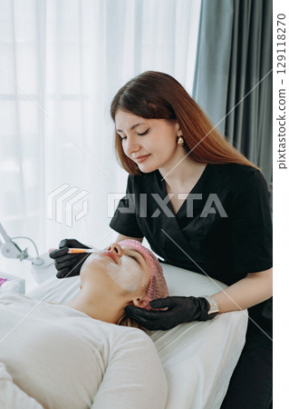 professional aesthetician applies a creamy face mask on young woman face using a brush professional aesthetician applies a creamy face mask on young woman face using a brush 129118270