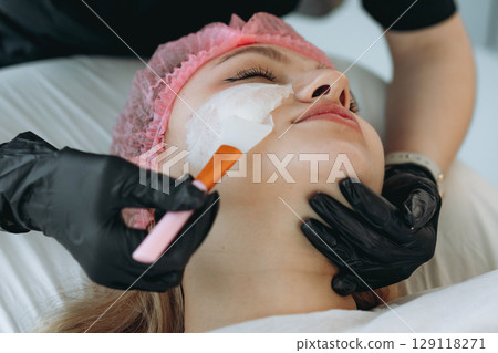 professional cosmetologist applies a creamy face mask on yiung woman face using a brush professional cosmetologist applies a creamy face mask on yiung woman face using a brush 129118271