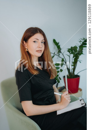 young woman dressed in black medical suit holding a clipboard seated on armchair in modern clinic young woman dressed in black medical suit holding a clipboard seated on armchair in modern clinic 129118869