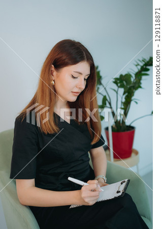 young woman dressed in black medical suit holding a clipboard seated on armchair in modern clinic 129118871