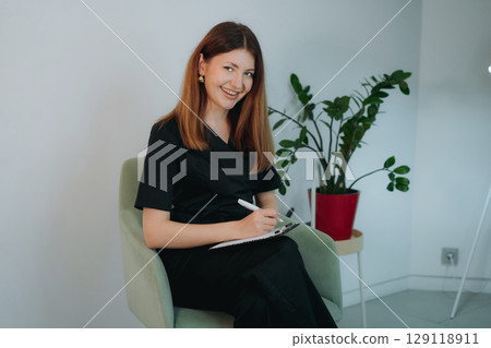young woman dressed in black medical suit holding a clipboard seated on armchair in modern clinic young woman dressed in black medical suit holding a clipboard seated on armchair in modern clinic 129118911