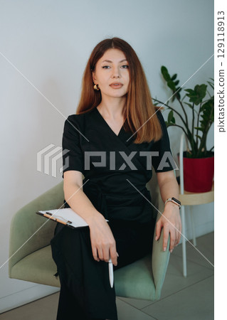 young woman dressed in black medical suit holding a clipboard seated on armchair in modern clinic 129118913