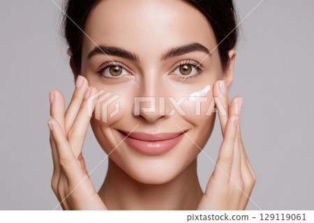 portrait of young beautiful woman with smooth healthy skin spreading a peptide-infused cream on her cheeks 129119061