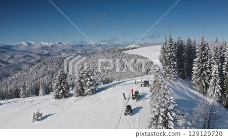 Aerial drone tourists riding snowmobiles and taking pictures in a snowy mountain forest, enjoying a breathtaking winter landscape on a sunny day. Bukovel ski resort. Winter travel background 129120720