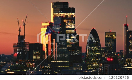 Warm sunset over London iconic skyline, featuring 30 St Mary Axe (The Gherkin), with construction cranes symbolizing growth in the bustling metropolis. Cityscape night background. Drone flight 129120736