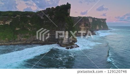 Aerial drone captures the breathtaking Uluwatu Temple perched atop a majestic cliff as waves crash against the rocks, creating a mesmerizing scene during a vibrant sunset 129120746