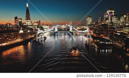 United Kingdom, London: Breathtaking aerial view of London at night, illuminated Tower Bridge, The Shard, and cityscape reflecting on the River Thames with boat, creating mesmerizing urban nightscape United Kingdom, London: Breathtaking aerial view of London at night, illuminated Tower Bridge, The Shard, and cityscape reflecting on the River Thames with boat, creating mesmerizing urban nightscape 129120778