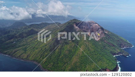Aerial drone showing smoke rising from the crater of Mount Iliwerung, an active volcano located on Flores Island, Indonesia, surrounded by lush green vegetation and the blue ocean 129120802