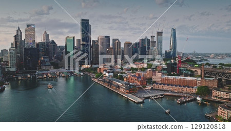 Australia, Sydney: CBD skyline reflecting on calm water in cloudy sunset, modern buildings cityscape skyscrapers towering over ferry cruises sailing ocean harbor. Aerial view drone panorama 129120818