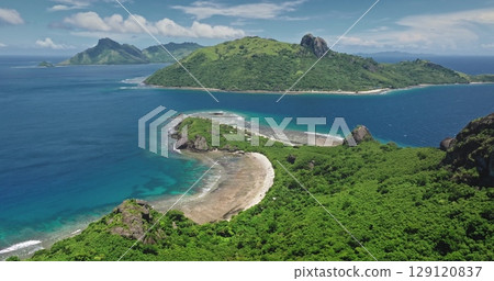 Lush tropical vegetation blankets the mountainous beach coastline terrain of Kuata Island, Fiji, surrounded by vibrant, crystal-clear waters beneath a bright blue sky. Drone flight 129120837