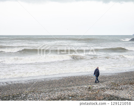 A lonely man walks along the shore of a stormy sea. Big waves on the sea. A man is walking on the coast. A lonely man walks along the shore of a stormy sea. Big waves on the sea. A man is walking on the coast. 129121058