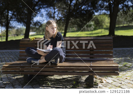 Young student drawing in notebook while sitting on park bench 129121186