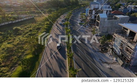 An aerial shot of a multi-lane road lined with palm trees and a mix of completed and under-construction buildings, with lush green fields on the left. The sun casts long shadows, suggesting late after An aerial shot of a multi-lane road lined with palm trees and a mix of completed and under-construction buildings, with lush green fields on the left. The sun casts long shadows, suggesting late after 129125320