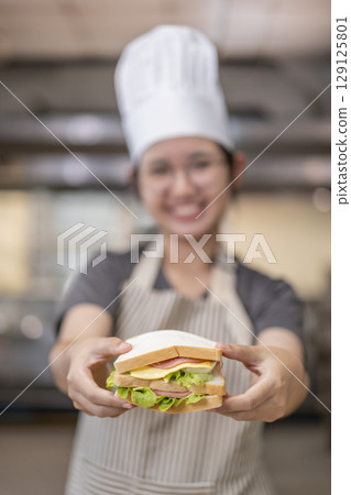 Smiling Asian Female Chef Holding Sandwich in Kitchen 129125801