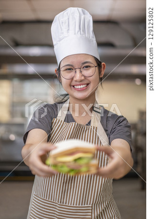 Smiling Asian Female Chef Holding Sandwich in Kitchen 129125802