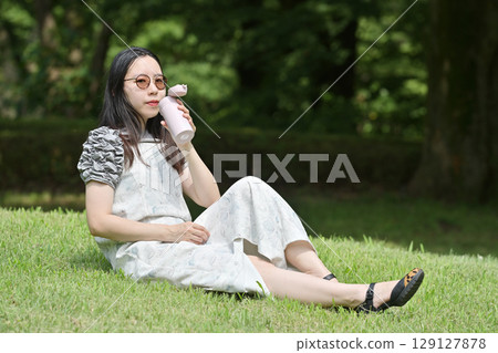 一位年輕女子坐在草地上，用自己的瓶子喝水 129127878