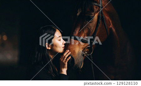 A young woman gently kisses a horse's face at sunset, capturing an intimate moment of trust and connection. The soft backlight highlights their bond in a serene countryside setting 129128110