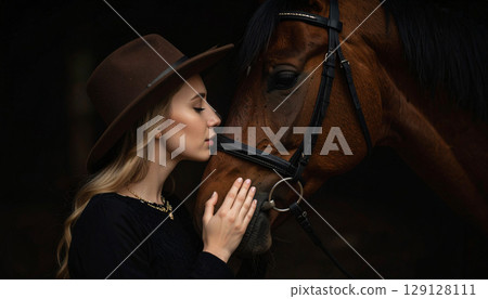 A young woman gently kisses a horse's face at sunset, capturing an intimate moment of trust and connection. The soft backlight highlights their bond in a serene countryside setting 129128111