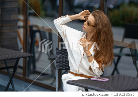 Outdoor portrait of a young stylish Caucasian woman sitting in sidewalk cafe. Vacation summer mood. Outdoor portrait of a young stylish Caucasian woman sitting in sidewalk cafe. Vacation summer mood. 129128555