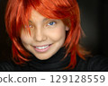 Close-up portrait of a red-haired smiley Caucasian teenager girl on a dark background. Fashion portrait of a little stylish female model. Photo with copy space. 129128559