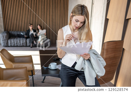 Businesswoman reading documents while thinking in office Focused businesswoman reading papers with two colleagues in background on a sofa Businesswoman reading documents while thinking in office Focused businesswoman reading papers with two colleagues in background on a sofa 129128574