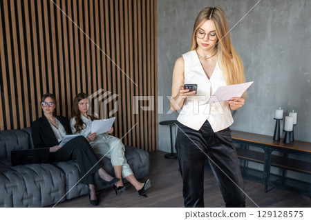 Businesswomen reviewing documents and phone in modern office lounge area. Businesswomen reviewing documents and phone in modern office lounge area. 129128575