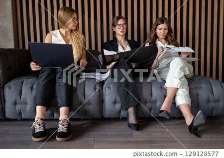Businesswomen reviewing project data on paper and laptop while seated on office couch Businesswomen reviewing project data on paper and laptop while seated on office couch 129128577
