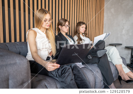 Businesswomen working together on couch, one using laptop while others read documents 129128579