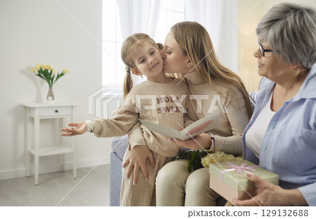 Child girl congratulating mother and grandmother on holiday sitting on sofa at home. 129132688