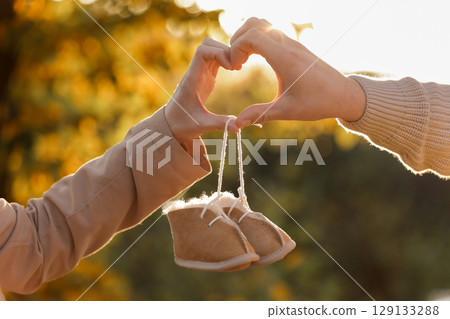 Man and pregnant wife are doing heart gesture with hands and holding warm baby shoes on nature autumn background. Future parents waiting for the baby. The concept of Mother's Day and Women's Day. 129133288
