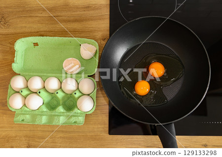 White chicken eggs in a cardboard egg tray on tabletop. Two raw eggs on frying pan on induction stove ready to cook. Delicious healthy simple breakfast meal. Natural egg protein to the diet. 129133298