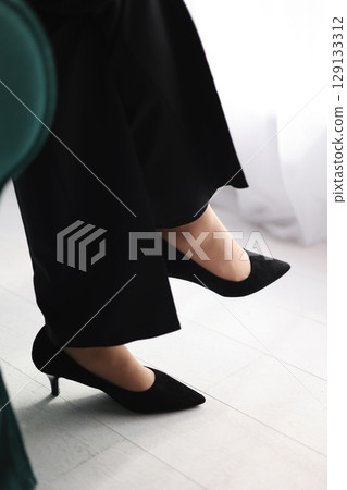 Cropped photo of legs in a classic black high heels shoes and black pants of a business woman sitting on a green chair by the window in the office. Cropped photo of legs in a classic black high heels shoes and black pants of a business woman sitting on a green chair by the window in the office. 129133312