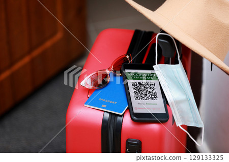 New normal concept. Close up view of a smartphone with an immune digital health passport, a red suitcase, protective mask, straw hat, passport and sunglasses. Traveling by plane during a pandemic. 129133325