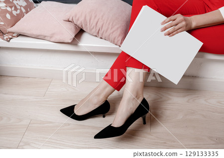 cropped photo of woman hands holding a magazine with mock up. girl in red pants and high heeled shoes sitting on sofa cropped photo of woman hands holding a magazine with mock up. girl in red pants and high heeled shoes sitting on sofa 129133335
