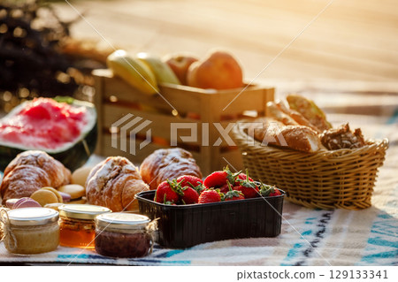 summer picnic basket with fruit and bakery on a blanket outdoors. Lunch with croissants, jam, watermelon, strawberry and fresh fruits in wooden box in the park. Copy space. summer picnic basket with fruit and bakery on a blanket outdoors. Lunch with croissants, jam, watermelon, strawberry and fresh fruits in wooden box in the park. Copy space. 129133341