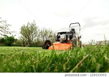 Gardening work tools. Wide angle view of orange electric lawn mower, wheels, motor on bright lush green grass at backyard. Rotary lawn mower machine cut lawn. Professional lawn care service. Gardening work tools. Wide angle view of orange electric lawn mower, wheels, motor on bright lush green grass at backyard. Rotary lawn mower machine cut lawn. Professional lawn care service. 129133353
