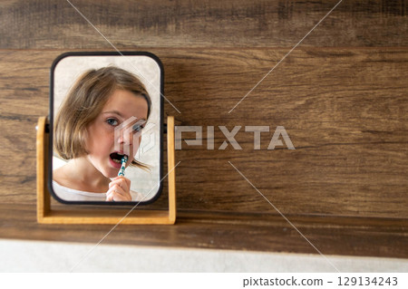 Oral hygiene, healthy teeth and care. Girl brushing teeth with toothbrush and looking in mirror in bathroom interior in the morning, closeup, empty space. High quality photo 129134243