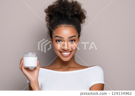 Beauty portrait of a young woman with a neat hairstyle with glowing skin holding a jar of cream on a neutral beige background 129134962