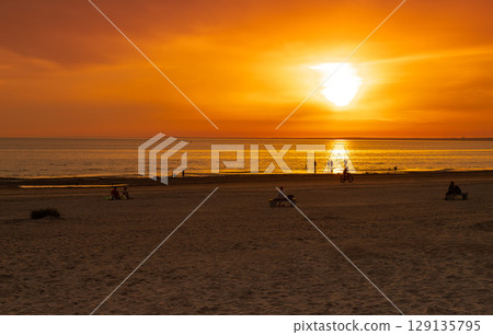 Summer evening at the beach. People watching the sunset over the sea. The sun sets on the water. The sky is painted with golden bright colors. Nature background. 129135795