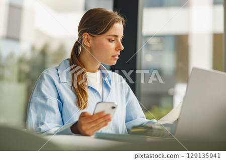 A focused young woman is diligently working on her laptop while simultaneously using her smartphone A focused young woman is diligently working on her laptop while simultaneously using her smartphone 129135941