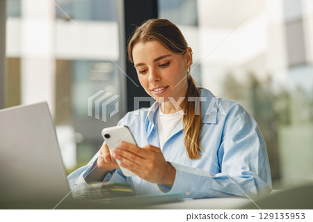 A Young Woman Utilizing Her Smartphone While Actively Working On Her Laptop Computer A Young Woman Utilizing Her Smartphone While Actively Working On Her Laptop Computer 129135953
