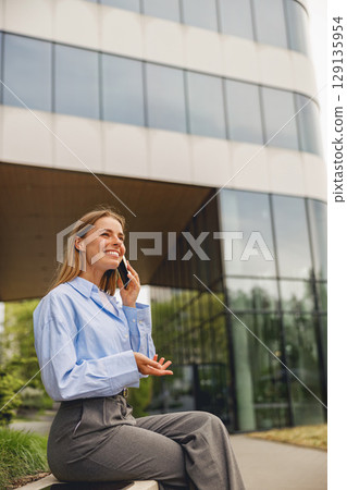 A businesswoman speaks on her phone outside a modern office, discussing important matters 129135954