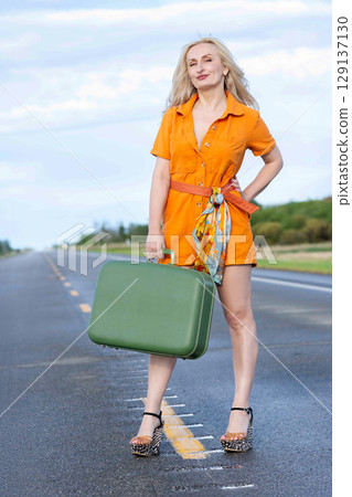 Blonde woman with a green retro suitcase standing on the highway. Blonde woman with a green retro suitcase standing on the highway. 129137130