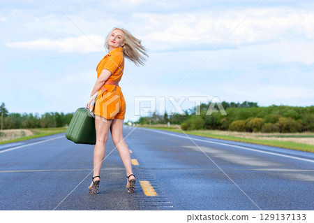 Blonde woman with a green retro suitcase standing on the highway. Blonde woman with a green retro suitcase standing on the highway. 129137133