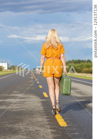 Blonde woman with a green retro suitcase walking on the highway. 129137136