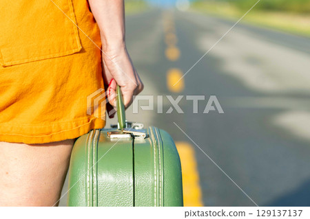 Woman's hand is holding a retro green suitcase and walking on a highway. Woman's hand is holding a retro green suitcase and walking on a highway. 129137137