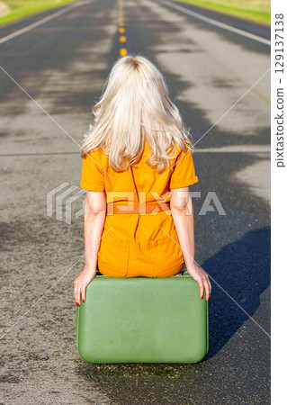 Blonde woman is sitting on a retro suitcase on a rural highway. Blonde woman is sitting on a retro suitcase on a rural highway. 129137138